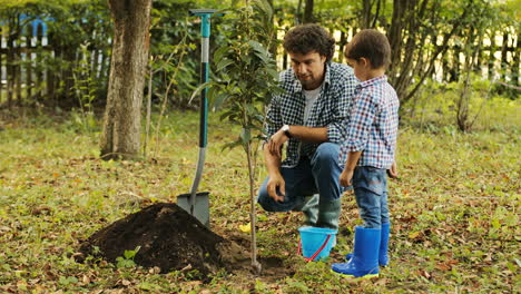 Portrait-of-a-little-boy-and-his-dad-planting-a-tree.-Dad-explains-something-to-his-son-and-puts-the-soil-with-the-toyspade-into-the-toybucket.-They-talk-and-smile.-Blurred-background